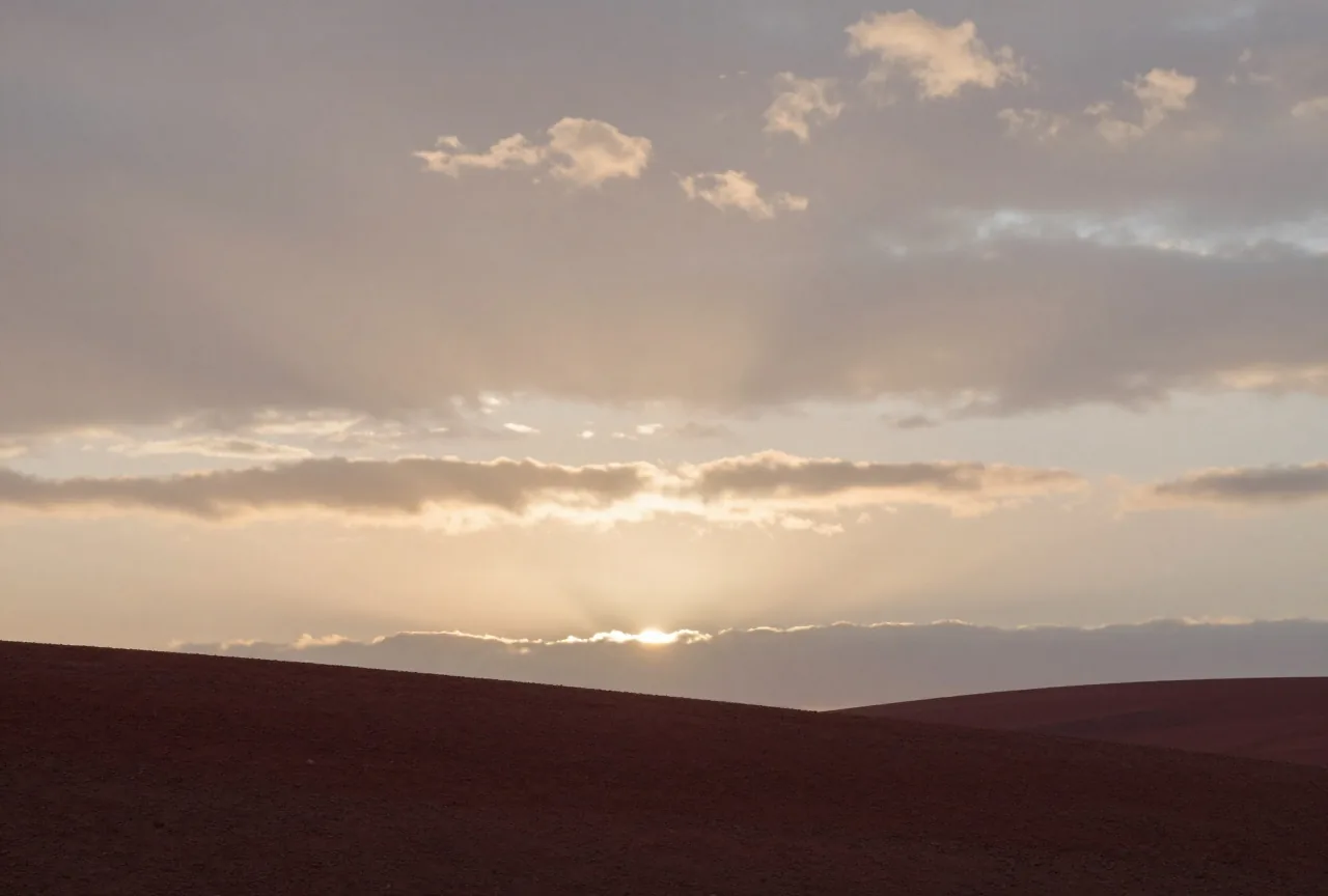 Ein stiller Hang bei Sonnenaufgang unter weitem Himmel mit Wolkenschichten, durch die Lichtstrahlen brechen