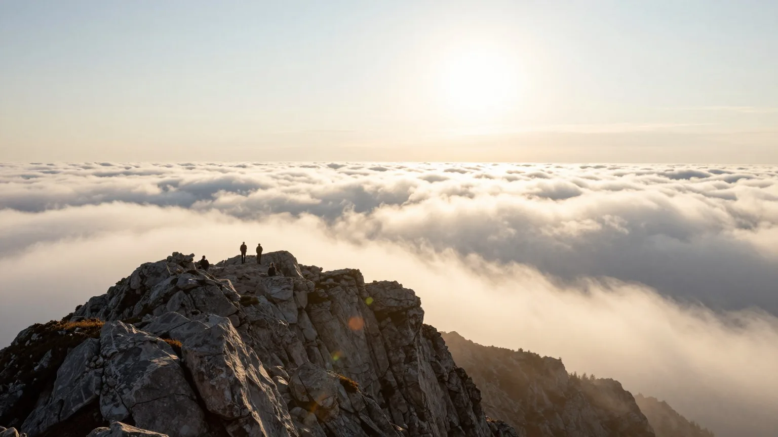 Ein felsiger Berggipfel über einem Wolkenmeer bei Sonnenaufgang, helles Licht bricht über den Grat