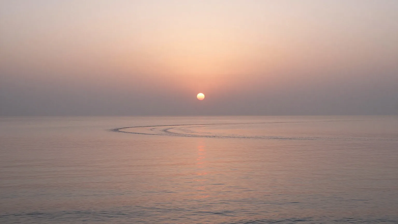 Sonnenaufgang über einem ruhigen Meer, in das ein Fluss mündet, Dunst liegt über dem Wasser