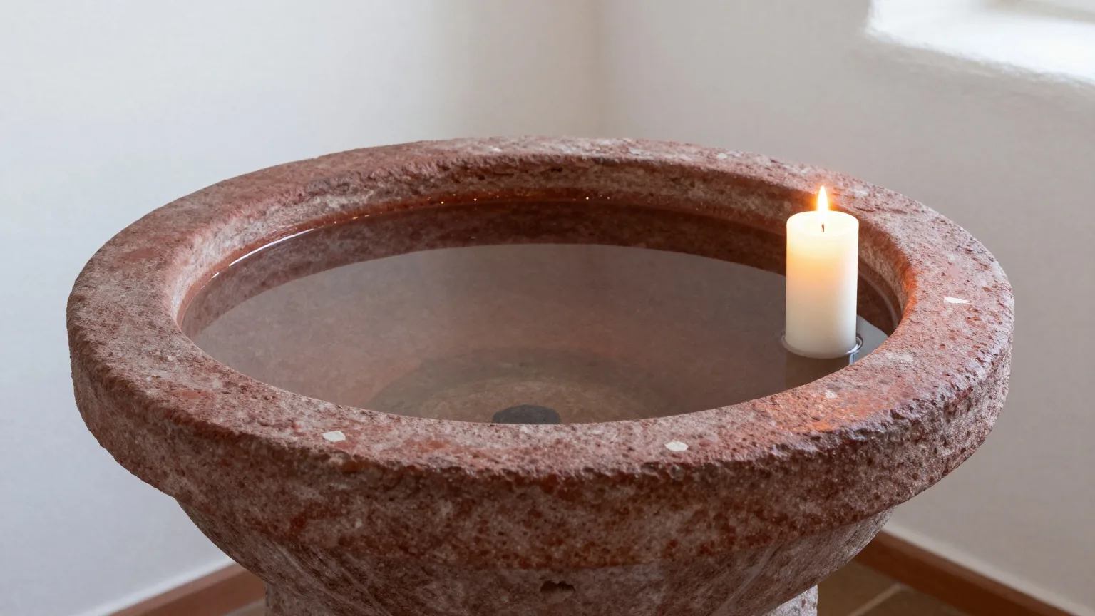 Ein schlichtes steinernes Taufbecken mit klarem Wasser in einer hellen weiß gekalkten Dorfkirche, daneben brennt eine weiße Kerze